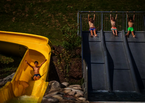 Ein Kind rutscht mit erhobenen Händen auf einer gelben Wasserrutsche, daneben halten sich drei Kinder an der Sicherheitsstange einer Trio-Slide fest.