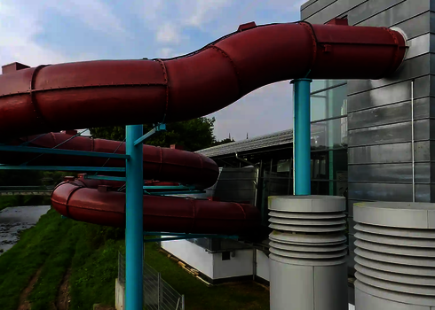 Gedämmte rote Wasserrutsche mit blauen Stützen verläuft um ein modernes Schwimmbadgebäude.