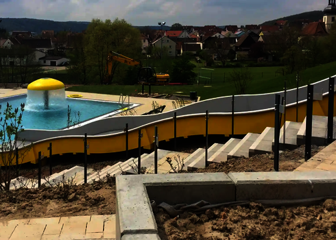 Blick auf eine gelbe Breitwellenrutsche, im Hintergrund ein Schwimmbecken und Häuser.