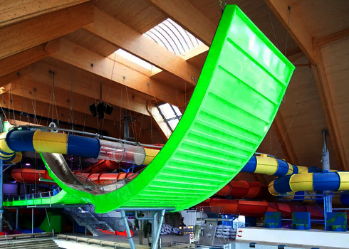 Grün leuchtende Boomerang-Rutsche im Innenraum, zwischen bunten Röhrenrutschen montiert.