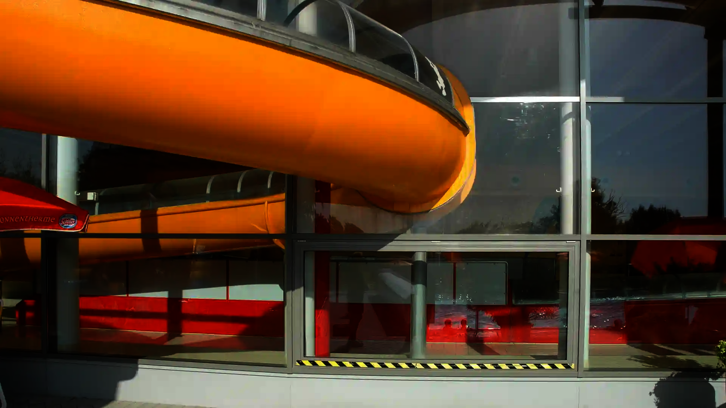 Gelbe Wasserrutsche durchquert eine Glasfassade und führt ins Freie.