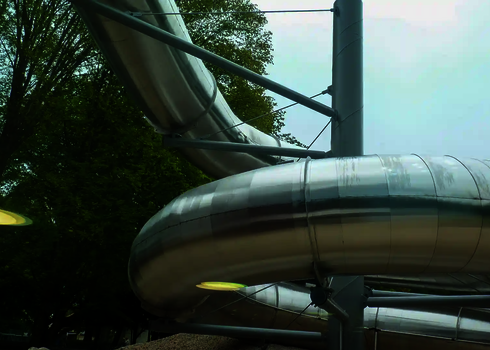 Abschnitt einer Edelstahl Wasserrutsche und Stahlstützen, vor Bäumen.
