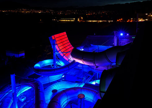 Boomerang-Rutsche bei Nacht mit bunter Beleuchtung in Blau und Rot vor Stadt-Panorama.