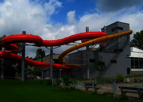  Rote und gelbe Wasserrutschen, die aus einem Gebäude herauskommen in parkähnlicher Umgebung.