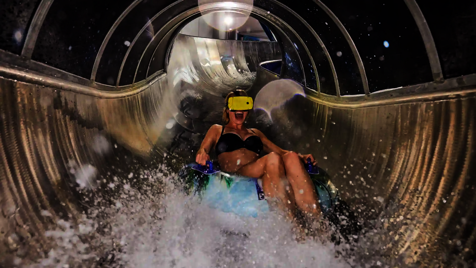 Frau mit gelber Virtual-Reality-Brille rutscht lachend auf einer Wasserrutsche.