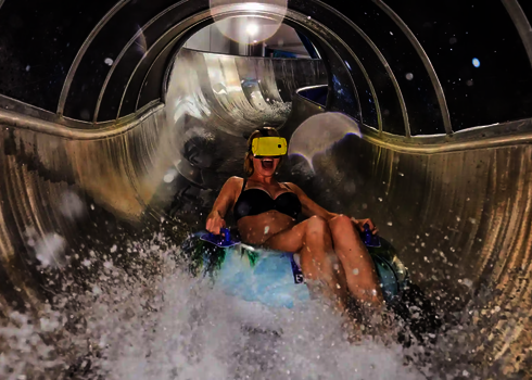 Frau mit gelber Virtual-Reality-Brille rutscht lachend auf einer Wasserrutsche.