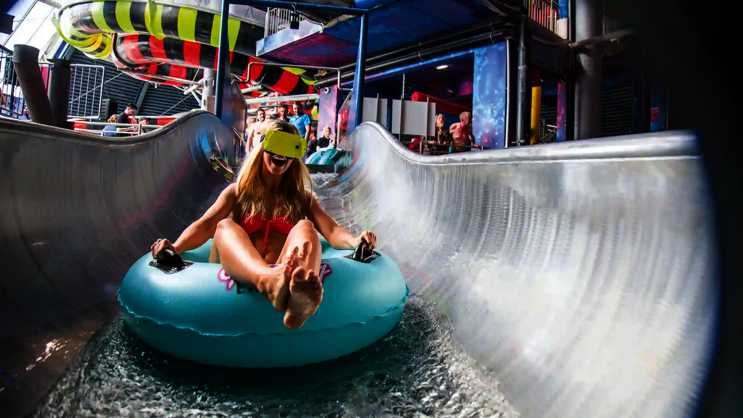 Eine blonde Frau sitzt lachend in einem blauen Rutschreifen, mit VR-Brille in einem Wasserpark.