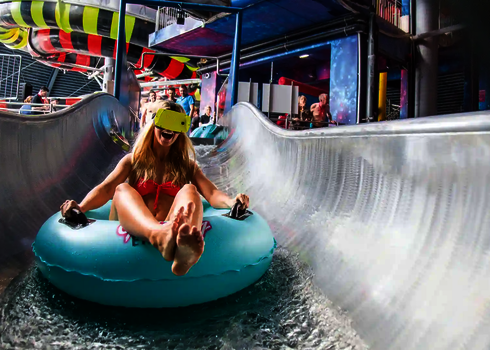 Eine blonde Frau sitzt lachend in einem blauen Rutschreifen, mit VR-Brille in einem Wasserpark.