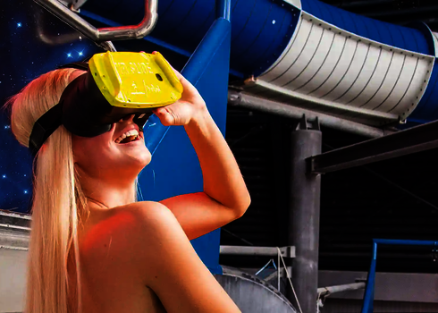 Blonde Frau trägt lachend eine gelbe VR-Brille, im Hintergrund Sternenbilder und eine Wasserrutsche.
