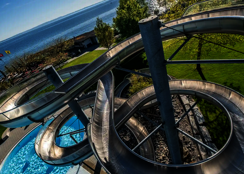 Mehrere Edelstahlröhrenrutschen winden sich über Felsen in Richtung Pool am Seeufer.