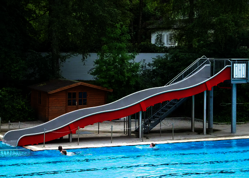 Eine lange Breitwellenrutsche mit roten Seitenwänden neben einem blauen Schwimmbecken.