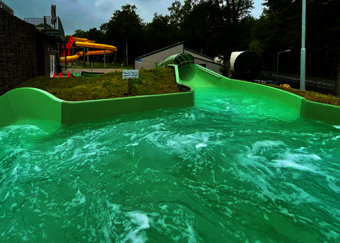 Grünes Becken mit kleinem Hinweisschild und gelber Röhrenrutsche im Hintergrund.