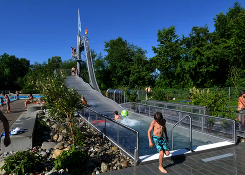 Children slide into a wide water slide with transparent side walls in the outdoor pool.