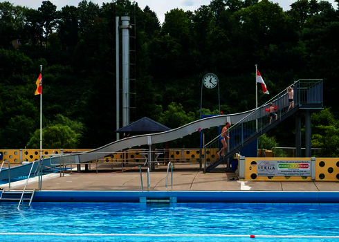 Seitenansicht einer Breitwellenrutsche, im Vordergrund ein Schwimmbecken, im Hintergrund Bäume.