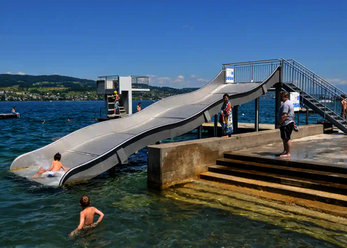 Eine Breitwellenrutsche steht in einem Badesee, drumherum befinden sich Menschen im Wasser oder an Land.