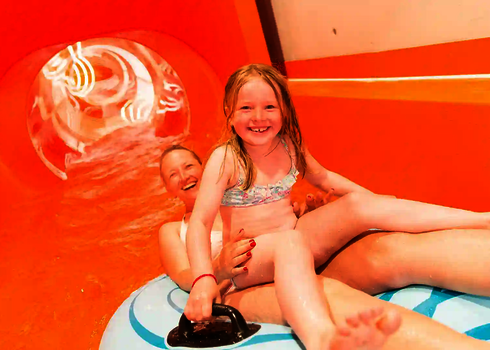 Kind sitzt lachend auf dem Schoß der Mutter, beide in einem Rutschreifen in einer Rutsche.