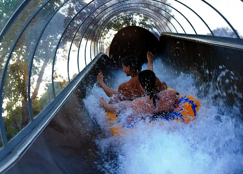 Zwei Personen rutschen auf einem gelben Doppelreifen durch einen transparenten Tunnel voller Spritzwasser.