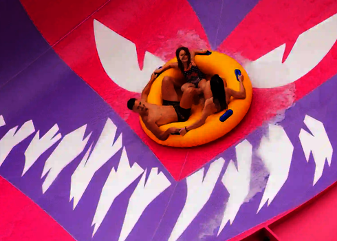 Drei Personen rutschen auf gelbem Rutschreifen durch die pinke Lotus Wasserrutsche.