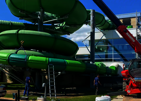 Grüne Röhrenrutsche mit Dämmung wird mit Kran montiert, Bauarbeiten sind im Gange.