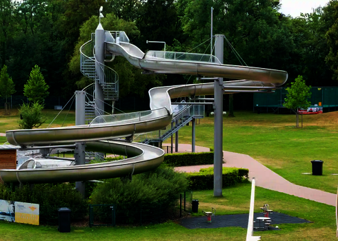 WR1040-Rutschenturm aus Edelstahl mit spiralförmigem Verlauf, zentralem Treppenturm und grüner Umgebung.