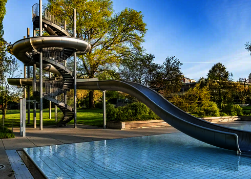 Eine Edelstahl-Körperrutsche windet sich in ein großes Freibadbecken, umgeben von Bäumen.