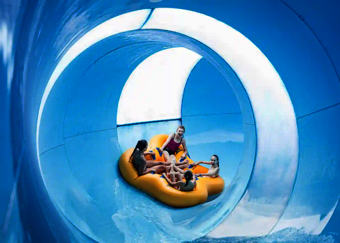 Vier Kinder rutschen lachend in einem gelben Rutschreifen durch eine blaue Röhrenrutsche.