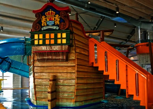 Hallenbad in dem ein Spielschiff für Kinder steht, von dem Wasserrutschen und eine Treppe angehen.