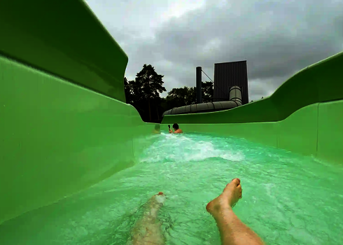 Zwei Personen treiben im Wildwasserfluss mit grüner Rutschbahn durch eine Kurve.