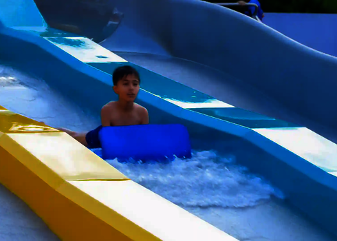 Child sliding down a two-coloured water slide in an amusement park with a blue slide mat.