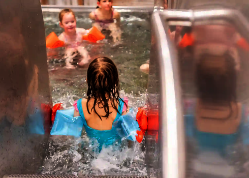  Kind mit Schwimmflügeln rutscht ins Wasser, andere Kinder warten im Becken.