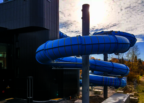 Blaue Outdoor-Wasserrutsche außerhalb eines Gebäudes mit gestreiften Tageslichteffekten.