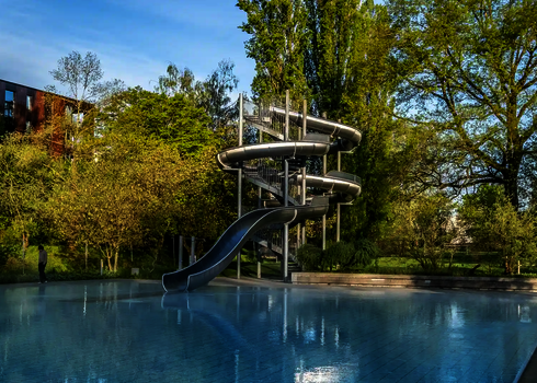 Eine Edelstahl-Körperrutsche windet sich in ein großes Freibadbecken, umgeben von Bäumen.