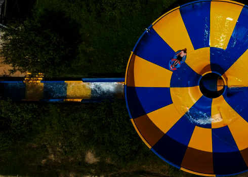 Blick von oben auf eine gelb-blaue Raft Bowl Rutsche in grüner Umgebung.