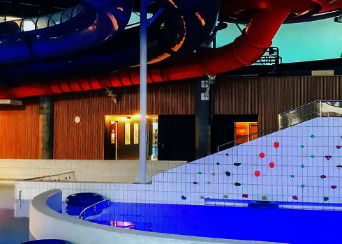 Innenansicht einen Hallenbads mit einem Wasserbecken und oben entlang verlaufenden Röhrenrutschen.