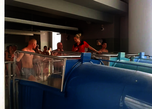 Badegäste stehen an blauen Röhrenrutschen an, eine Aufsichtsperson gibt Anweisungen.