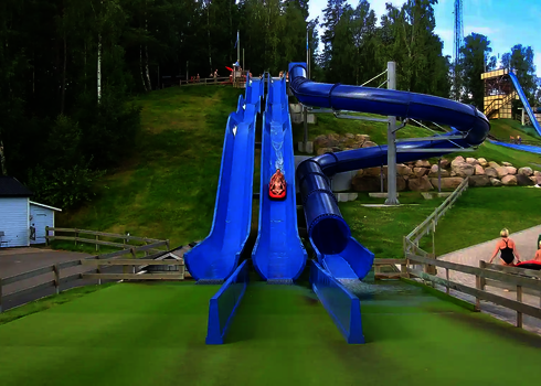 Blaue Wasserrutschen laufen einen grünen Berghang hinab, Personen sind am Rutschen.