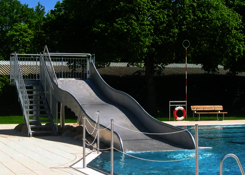Eine kurze Breitwellenrutsche mit einem Treppenaufgang, beides befindet sich neben einem Schwimmbecken, im Hintergrund Bäume.