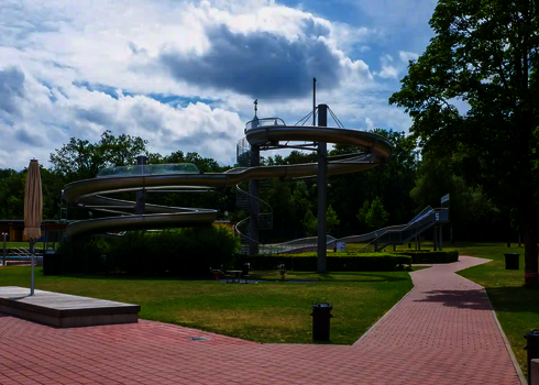 WR1040-Rutschenturm mit Doppelspirale in Edelstahloptik, aufgestellt in weitläufigem Freibadgelände.