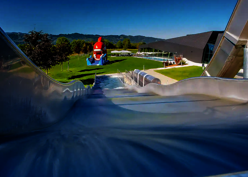 Sicht von oben auf eine Breitwellenrutsche, im Hintergrund einer bunter Kinderspielbereich auf einer grünen Wiese.