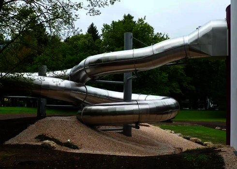 Geschwungene Edelstahlrutsche mit Fassadendurchdringung in parkähnlicher Umgebung. 