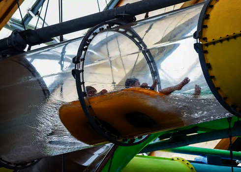 Menschen rutschen auf einem gelben Rutschreifen durch eine transparente Acrylröhre.