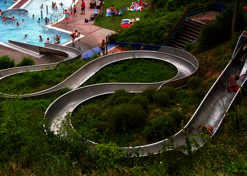 Eine gewundene Edelstahl-Wasserrutsche, im Hintergrund ein Schwimmbecken mit Badegästen.
