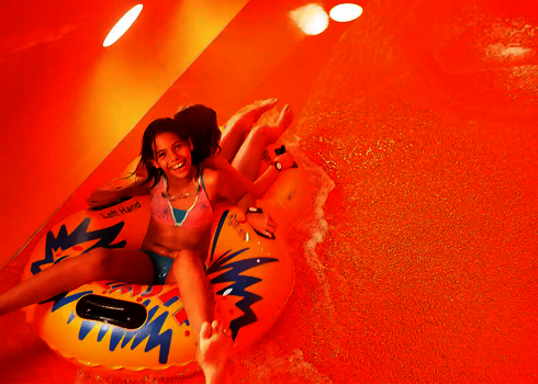 Zwei Kinder rutschen lachend auf einem Reifen durch eine orange beleuchtete Röhrenrutsche.