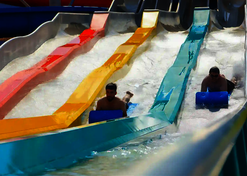 Two men slide down a colourful water slide in an outdoor pool using slide mats.