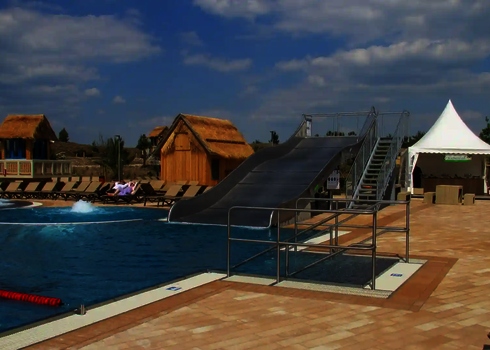 Eine Breitwellenrutsche mündet in ein Schwimmbecken, links stehen Strohhäuser und rechts ein weißes Zelt.