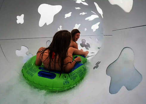 Innenansicht einer Röhrenrutsche in der zwei Frauen auf einem grünen Rutschreifen rutschen.