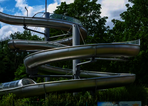 Gewundene Edelstahl Wasserrutsche in naturnaher Umgebung vor blauem Himmel.