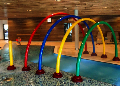Wasserspielplatz mit Wie-Spray Elementen "Regenbogen" und spielenden Kindern im Hintergrund.