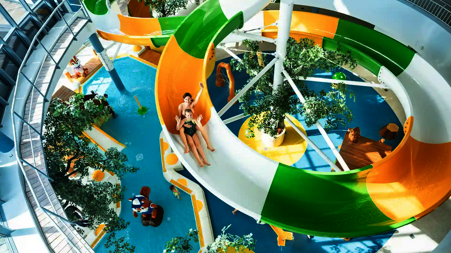 Zwei Personen rutschen eine bunte WR1040 Wasserrutsche in einem Schwimmbad herunter und winken in die Kamera.