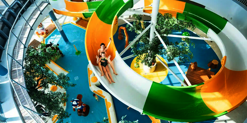 Zwei Personen rutschen eine bunte WR1040 Wasserrutsche in einem Schwimmbad herunter und winken in die Kamera.